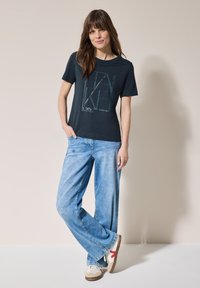 Marineblå t-shirt med grafisk print, parret med lyseblå wide-leg jeans. Sneakers har hvid base med røde accenter. Struktureret hår.