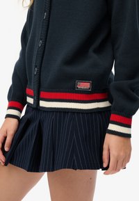 Cardigan en maille bleu marine avec une texture côtelée, présentant des accents à rayures rouges, blanches et bleu marine ainsi qu'un écusson. Associé à une jupe bleu marine à fines rayures.