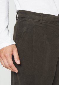 Hnědé kordurové kalhoty s žebrovanou texturou, s plisovaným předním dílem, vysokým pasem a bočními kapsami, doplněné bílou košilí s dlouhým rukávem.