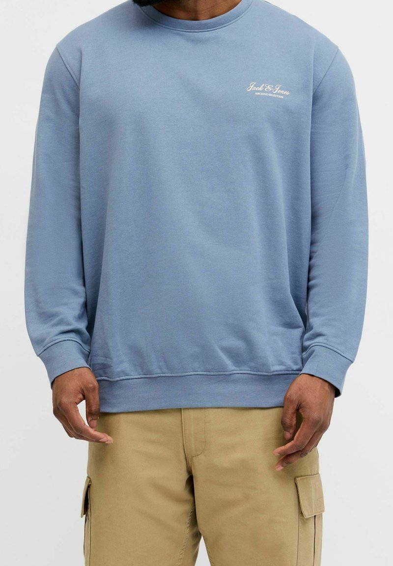 Homme portant un sweat-shirt bleu clair avec le logo "Jack & Jones" sur la poitrine et un pantalon cargo beige, debout devant un fond uni.