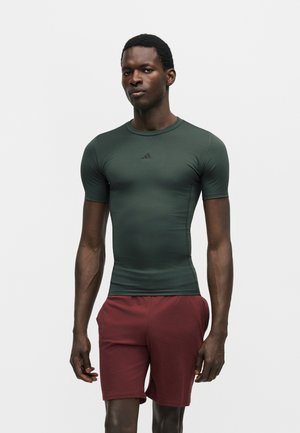 Homme debout portant un t-shirt de sport ajusté à manches courtes vert foncé et un short grenat sur un fond blanc uni.