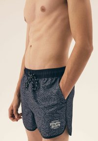 Niebieskie kąpielówki w wzory z paskiem ściągającym w talii, z białym napisem i teksturowanym wzorem, sięgające do połowy ud.