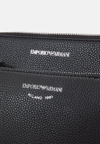 Černá texturovaná kožená peněženka s vyraženým stříbrným logem "EMPORIO ARMANI" a detailen "MILANO 1981". Zapínání na zip podél horního okraje.