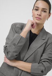 Grijze gestreepte blazer met gestructureerde schouders, brede revers en knopenmanchetten. Draagt over een zwarte top met een gladde textuur.