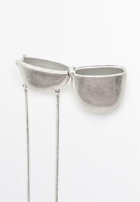 Zilveren metalen accessoire met twee ronde compartimenten die door een ketting met elkaar zijn verbonden. Gelaagd oppervlak met een gepolijste afwerking.