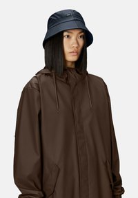 Giacca impermeabile marrone con cappuccio e coulisse, abbinata a un cappello a secchiello nero con una patch con logo sulla parte anteriore.