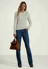 Ženska v belem puloverju z kabelskim vzorcem in modrih hlačah drži rjavo usnjeno torbico, stoji pred enobarvno ozadjem.