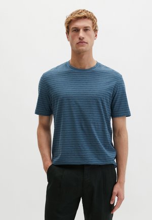 Mężczyzna o krótkich blond włosach, ubrany w niebieską koszulkę w paski z krótkim rękawem i okrągłym dekoltem oraz czarne spodnie, stoi na jednolitym białym tle.