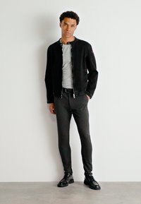 Veste bomber noire, t-shirt gris, pantalon sombre ajusté, chaussures noires. Texture lisse avec des poignets élastiques. Le mannequin pose devant un fond uni.