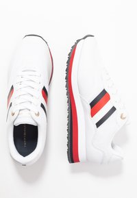 Baskets blanches avec des rayures rouges et noires, des lacets blancs et des semelles noir et rouge, présentées en vues de dessus et de côté.