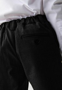 Schwarze Hose mit elastischem Bund, ausgestattet mit einer kleinen Hintertasche mit Knopf. Der Stoff wirkt weich und strukturiert.