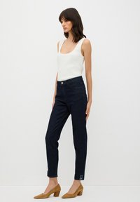 Jeans de talle alto azul oscuro con una forma ajustada, sutiles bolsillos delanteros y bordado floral decorativo en el dobladillo, combinados con una camiseta sin mangas blanca.