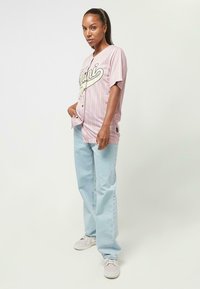 Rosa gestreiftes Jersey-Shirt mit schwarzen Knöpfen und grafischem Text, kombiniert mit hellblauen Jeans und hellen Sneakers, aufrecht stehend.