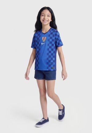 Osmijehnuta djevojka hoda, noseći plavi sportski dres na kockice, tamne kratke hlače i mornarsko plave tenisice, na jednostavnoj svijetloj pozadini.