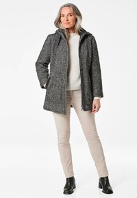 Houndstooth-gepatroneerde jas in grijs en zwart met een capuchon, ritsluiting, zijzakken en contrasterende zwarte accenten. Draag hem over een crème trui en beige broek.