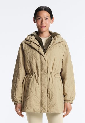 Femeie purtând o geacă matlasată bej cu glugă, cu talie și manșete elastice, suprapusă peste un pulover verde-olive cu fermoar.