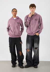 Μωβ φούτερ με φερμουάρ και λογότυπο μπροστά, φορεμένο με σκισμένα μαύρα και γκρι τζιν που έχουν δερμάτινες άκρες και τρύπες, σε συνδυασμό με μαύρα παπούτσια.