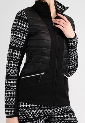 Frau trägt eine schwarze gesteppte Weste mit Reißverschlusstaschen über einem schwarz-weißen gemusterten Langarmshirt und Hose, Hand nahe am Reißverschluss des Kragens.