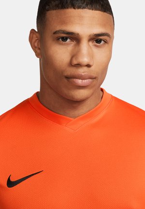 Nike orange Sport-Shirt mit V-Ausschnitt, strukturiertem Stoff und schwarzem Logo auf der linken Seite. Nahaufnahme des Halsausschnitts und oberen Brustbereichs.