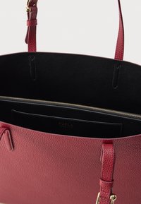 Sarkans graudainas ādas tote soma ar melnu iekšpusi, zelta detaļām un kabatu ar rāvējslēdzēju. Tai ir divas rokturu uznešanas iespējas.