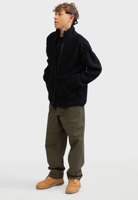 Black fleece jacket with a high collar, front zip, and two exterior pockets. Worn with olive green cargo pants and brown boots.