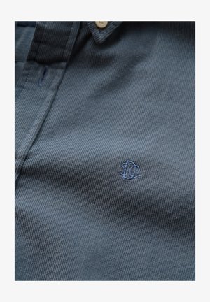 Zoom su una camicia di velluto a coste blu con un piccolo logo di pesce ricamato vicino al petto e un bottone beige vicino al colletto.