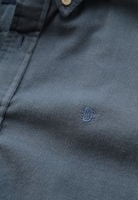 Dark gray corduroy shirt featuring a buttoned collar, vertical ribbed texture, and a small blue embroidered logo on the chest.