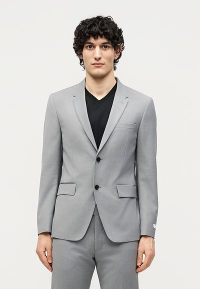 Νέος άνδρας με σγουρά μαλλιά που φοράει ανοιχτό γκρι κοστούμι και μαύρο μπλουζάκι με V-λαιμόκοψη, στέκεται μπροστά σε λευκό φόντο.