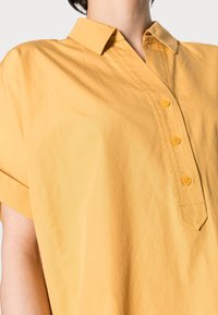 Blusa gialla a maniche corte con colletto, caratterizzata da una patta con quattro bottoni gialli e una vestibilità comoda. Texture liscia.