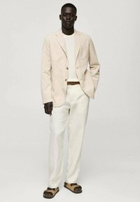 Blazer in lino beige chiaro con due bottoni, abbinato a un maglione bianco a coste e pantaloni off-white. Un cinturone marrone e sandali color cammello completano il look.
