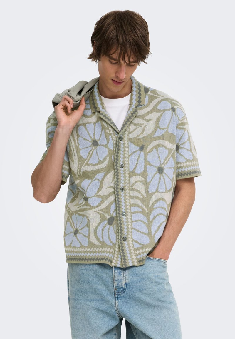 Jeune homme portant une chemise à manches courtes florale verte et bleue avec un jean bleu clair, tenant une veste grise sur son épaule.