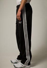 Czarne spodnie sportowe z trzema pionowymi białymi paskami, elastycznym pasem i haftowanym emblematem na lewej biodrze. Materiał wydaje się miękki.