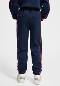 Mörkblå joggers med elastisk midja, kontrasterande röda sidostreck och elastiska muddar. Bärs med ljust färgade sportskor.