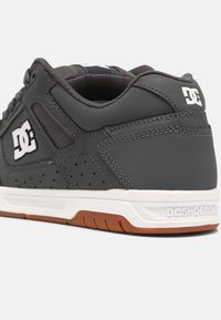 Gray sneaker with white accents, textured paneling, perforations, rubber outsole, and logo detail on the heel and side.