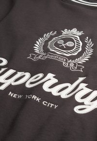 Dark gray sweatshirt with white embroidered text "Superdry" and "New York City." Features decorative emblem with tennis rackets and laurel.