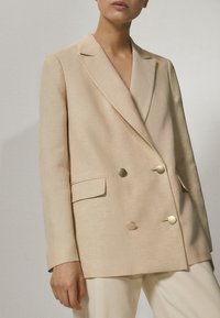 Beige, leinen, doppelt knöpfbarer Blazer mit zwei großen goldfarbene Knöpfen, Reverskragen und Seitentaschen. Glatte Textur und entspannte Passform.