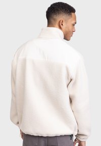 Fleece-takki off-white-värissä, jossa on korkea kaulus, teksturoitu vartalo ja sileä olkapaneli, varustettuna säädettävillä nyöreillä helman kohdalla.