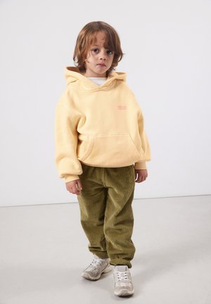 Young child with light brown hair wearing a yellow hoodie, olive green corduroy pants, and white sneakers against a plain background.