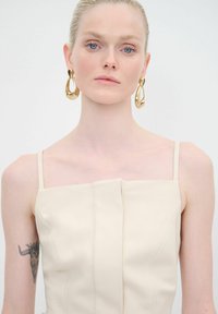 Haut sans manches beige avec un décolleté carré, présentant de doux plis et une délicate couture ; le mannequin porte de grandes boucles d'oreilles en or.