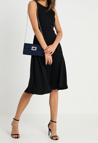 Žena v černých midi šatech bez rukávů a černých vysokých podpatcích, která drží modrou kabelku s ozdobnou sponou.