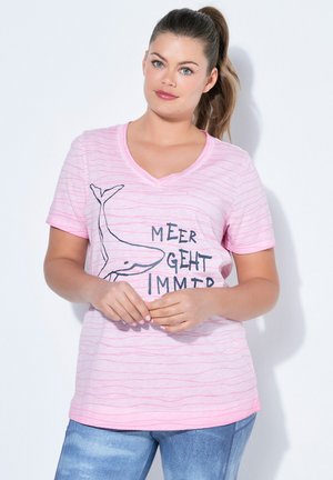 Kvinde med brunt hår i en hestehale iført en lyserød stribet V-hals T-shirt med en hvaltegning og tysk tekst, kombineret med blå jeans.