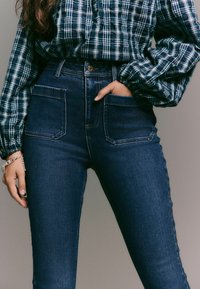 Pantalones vaqueros oscuros de cintura alta con un corte ajustado, que presentan grandes bolsillos frontales y detalles de costura sutiles. Combinados con una camisa de patrón a cuadros.