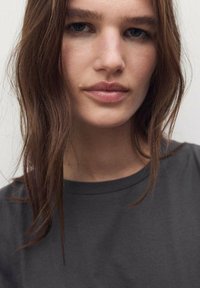 Detaljan prikaz osobe s dugom, smešnom kosom. Nose tamnupu polo majicu s okruglim izrezom. Prirodni make-up naglašava neutralan izraz lica.