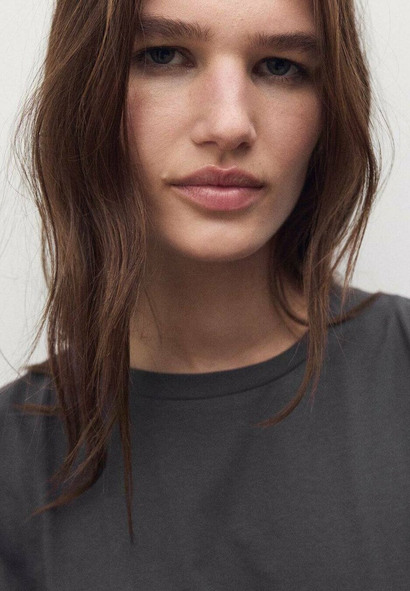 Detaljan prikaz osobe s dugom, smešnom kosom. Nose tamnupu polo majicu s okruglim izrezom. Prirodni make-up naglašava neutralan izraz lica.