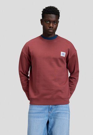 Hombre de pie mirando al frente, lleva una sudadera burdeos con un parche del logo de Lee y vaqueros azul claro, sobre un fondo gris claro liso.