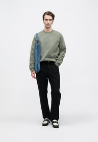 Mlad moški stoji v olivno zeleni majici, črnih hlačah, črno-belih čevljih in nosi modro torbo čez ramo.