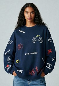 Marine sweatshirt met verschillende geborduurde ontwerpen, waaronder een roos, slangen, sterren en tekst. Beschikt over een geribbelde halslijn en een oversized pasvorm.
