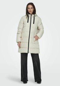 Manteau d'hiver long en crème avec capuche, doté d'une fermeture éclair frontale intégrale, de cordons noirs et de poches latérales. Texture douce et matelassée.