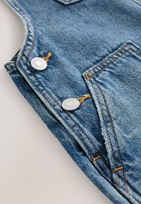 Salopette en denim bleu clair avec des coutures cuivre, dotée de deux boutons argentés et d'un tissu texturé. Détail remarquable avec une poche avant.