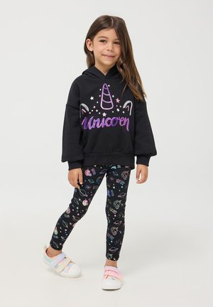 Czarna bluza z kapturem z napisem "Jednorożec" i grafiką, w połączeniu z czarnymi legginsami z kolorowymi wzorami inspirowanymi kosmosem; białe buty z różowymi paskami.